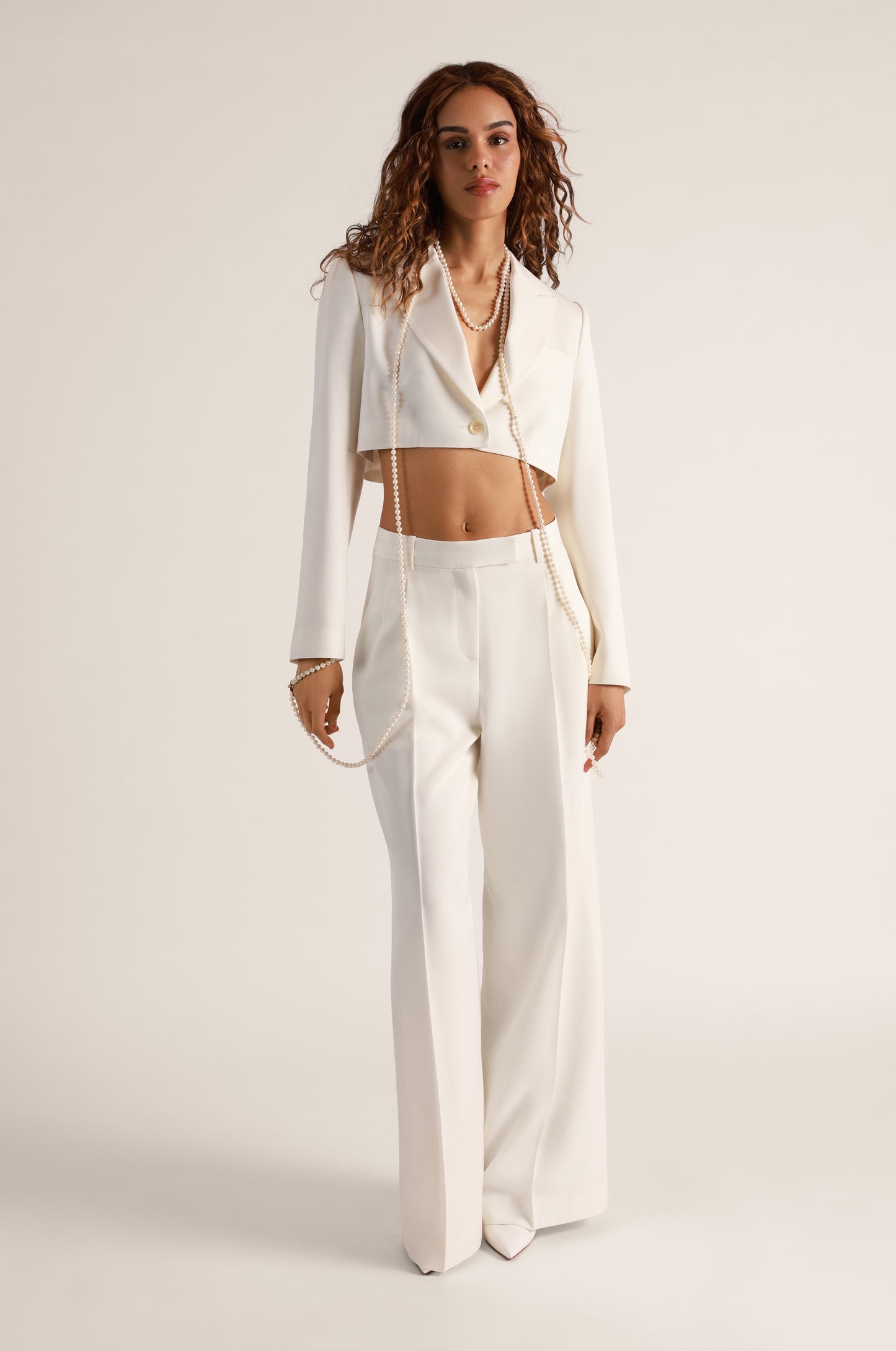 A female model wearing a white cropped blazer with long sleeves, paired with a matching suit tailored mini skirt and wide-leg double pleat pants.